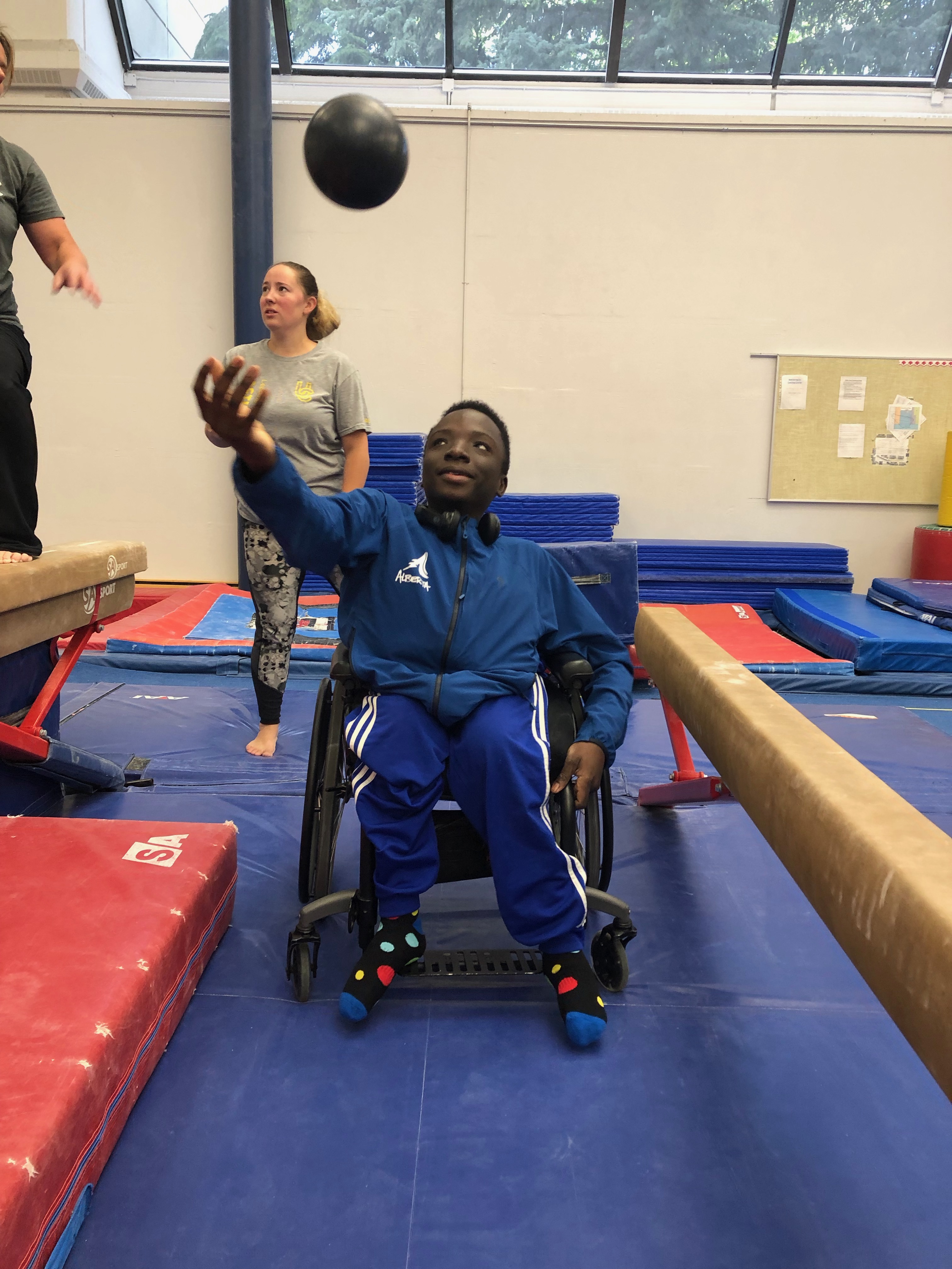 Jeune garçon en fauteuil roulant qui lance en l’air un ballon d’entraînement noir. Il est dans un gymnase.